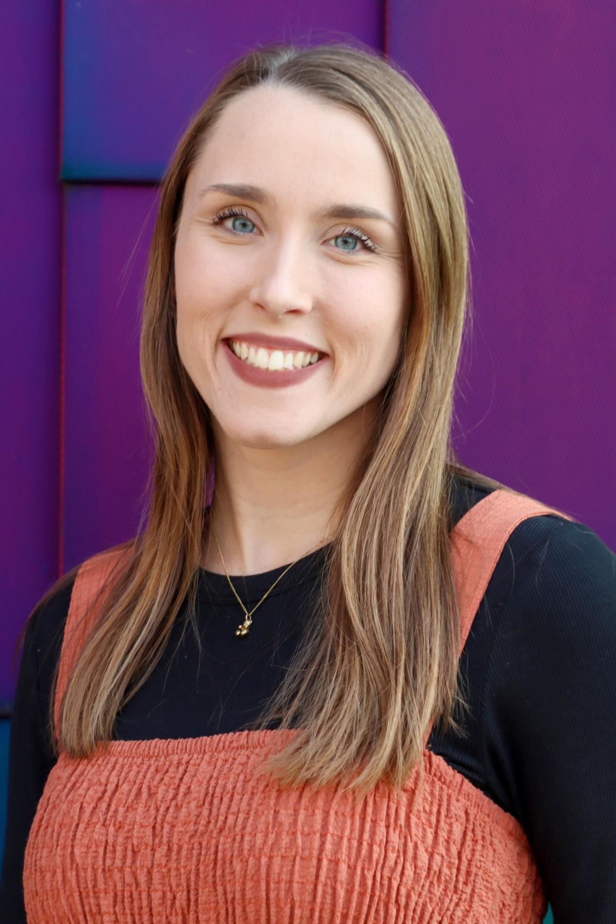 Professional headshot of Claire Sybesma, smiling and wearing a rust-orange dress over a black top, with a purple background