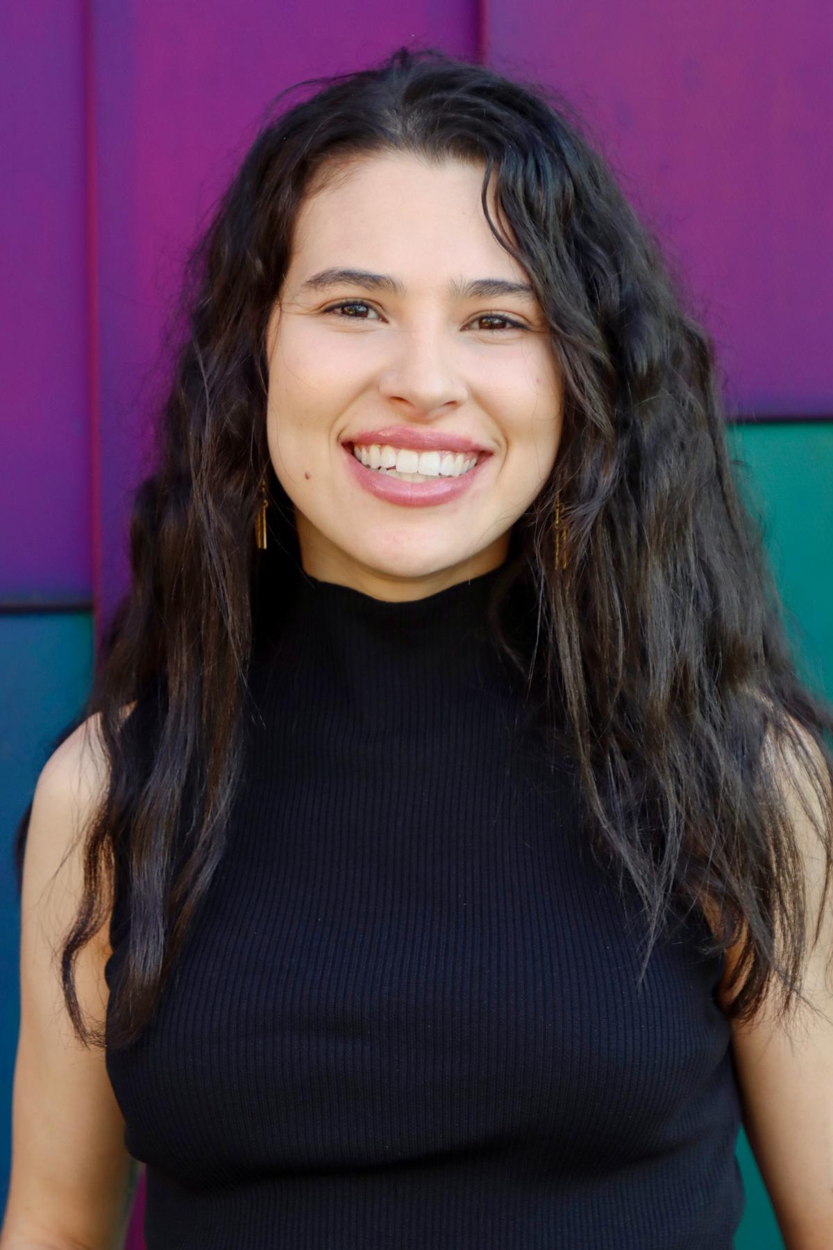 Isabella has long wavy dark hair, wearing a black sleeveless top, smiling in front of a colorful purple and teal background.