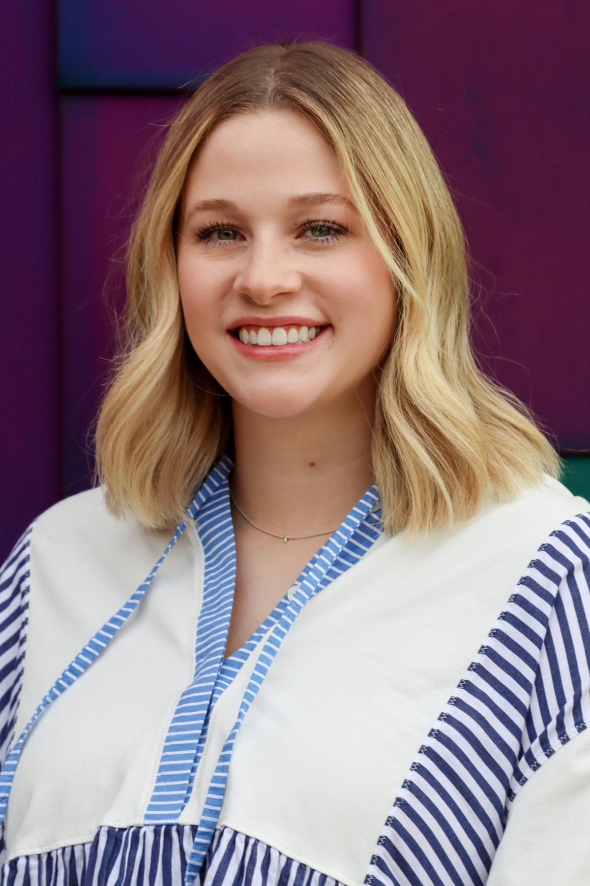 Smiling woman with shoulder-length blonde hair wearing a white and blue striped blouse, standing in front of a purple background.