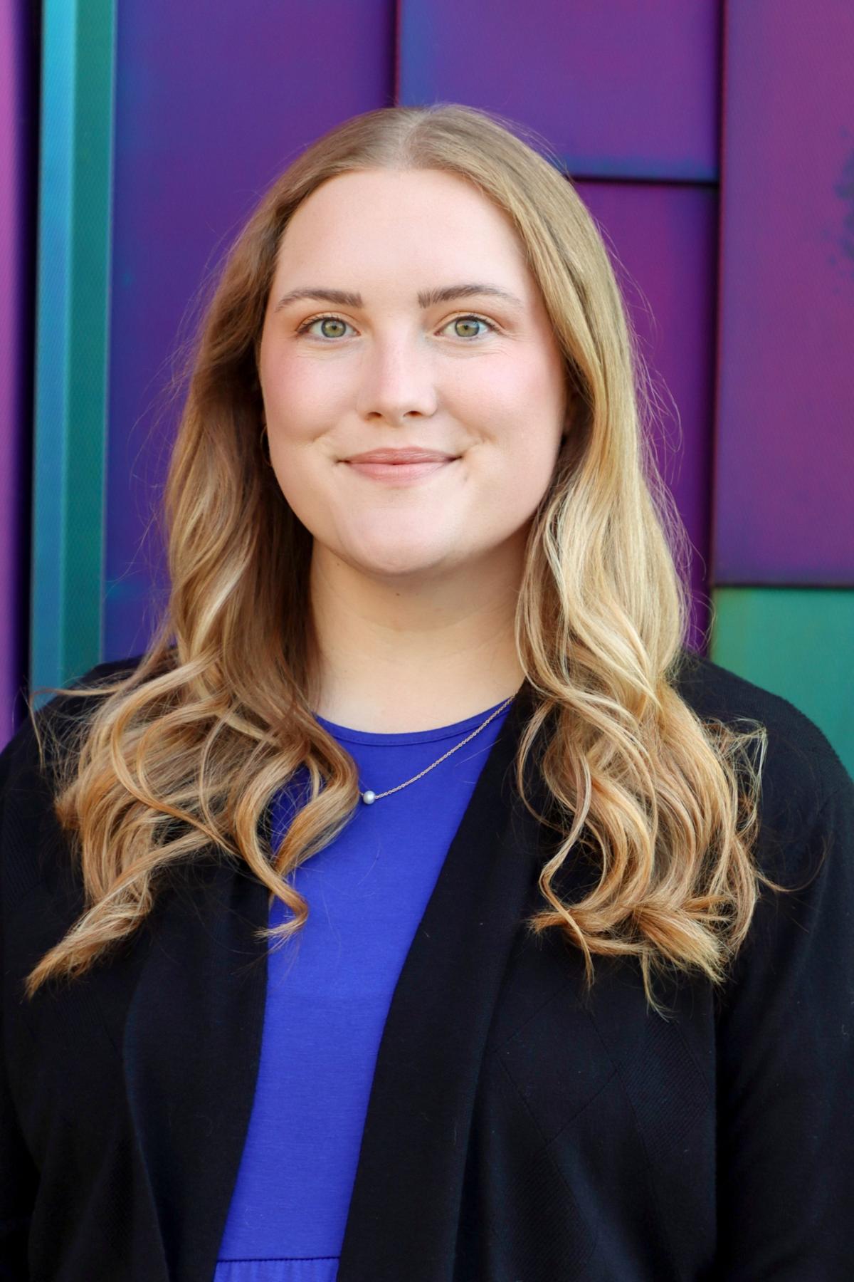 Professional headshot of a woman with long blonde hair, wearing a blue top and black cardigan, smiling in front of a colorful purple and teal background.