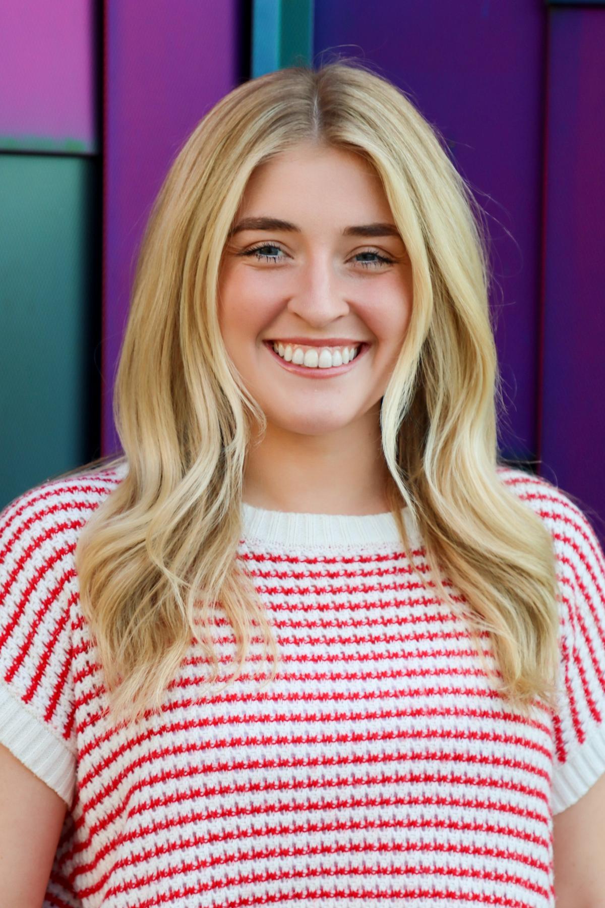Professional headshot of a woman with straight blonde hair, wearing a white sweater with red stripes, smiling in front of a colorful purple and teal background.