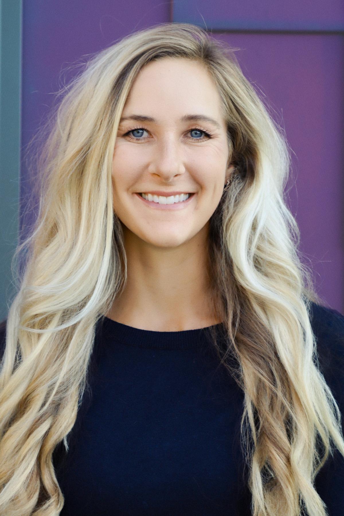 Professional headshot of a woman with long wavy blonde hair, wearing a black top, smiling in front of a colorful purple and teal background.