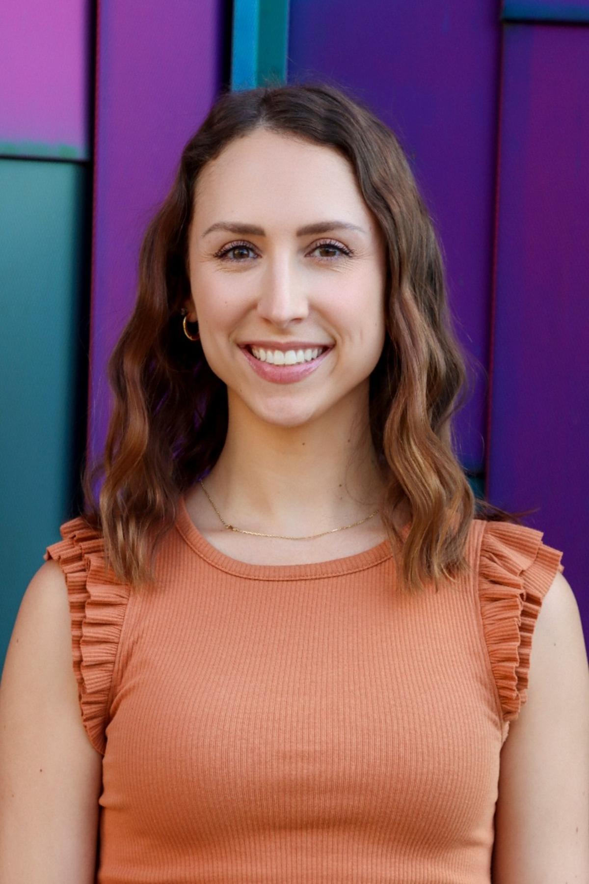 Professional headshot of a woman with shoulder-length wavy brown hair, wearing an orange sleeveless top, smiling in front of a colorful purple and teal background.